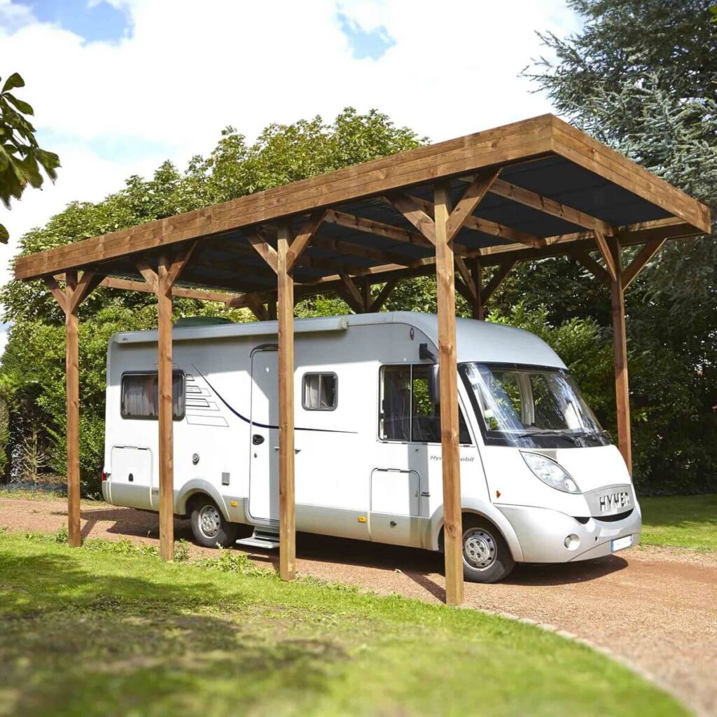 Carport en bois avec camping-car garé, sur allée gravillonnée entourée d'arbres